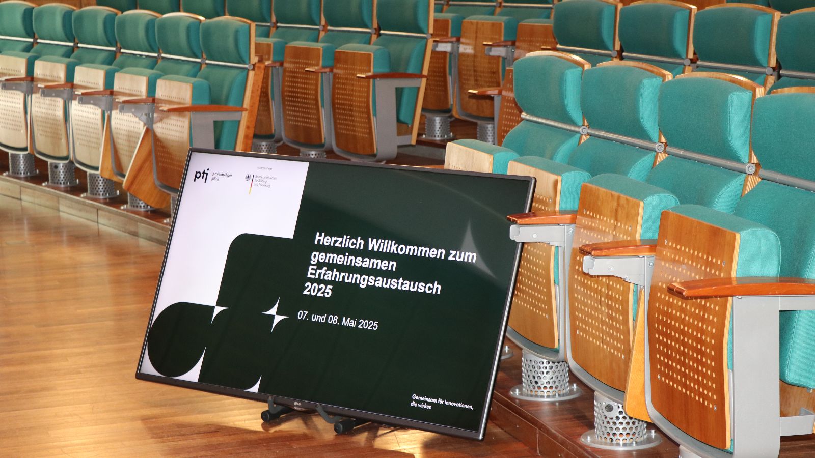 Screen vor Stuhlreihen im Hörsaal zeigt die Titelfolie der Präsentation zur Veranstaltung.