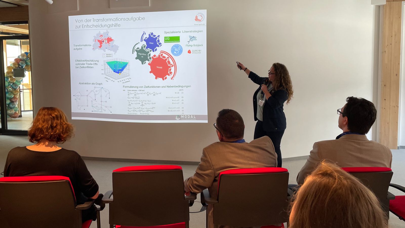Blick aus dem Publikum auf die Vortragende. In ihrem Rücken eine Präsentation auf einer Leinwand.